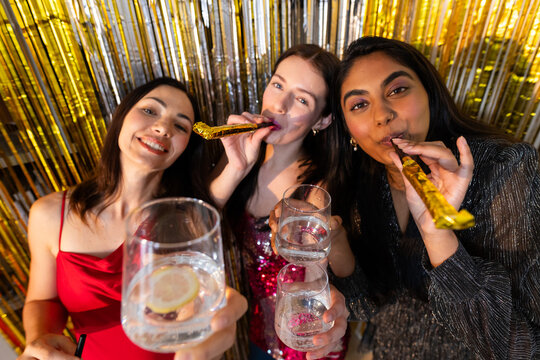 Diverse female trio in dresses posing at gold fringe, holding lemon glasses, blowing gold blowers