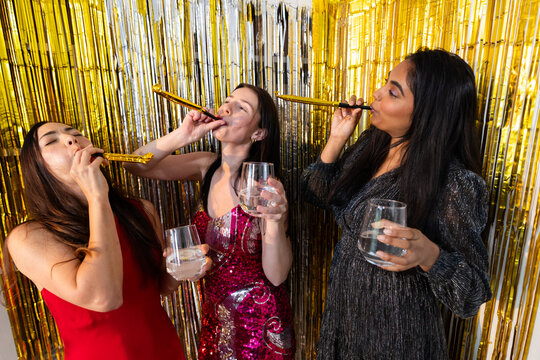 Diverse female friends in sequins blowing party horns and holding clear glasses at fringe backdrop