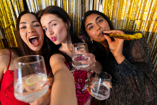 Diverse female friends taking selfie at gold fringe backdrop in sequins holding stemless glasses