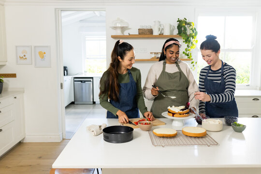 Diverse women friends assembling and frosting layered cake in kitchen using turntable