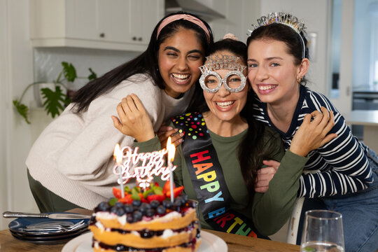 Three female friends celebrating birthday in home kitchen at wooden table with berry cake and sash