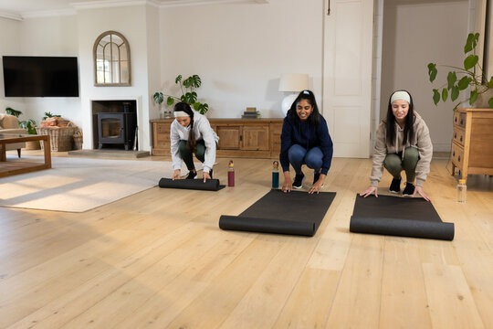 Diverse female trio in activewear kneeling and unrolling black yoga mats at home with water bottles