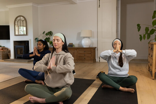 Diverse female friends practicing meditation on black yoga mats in living room, wearing headbands