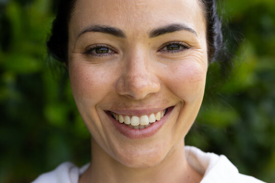 Adult female smiling, showing teeth and freckles, facing camera in garden with foliage, white top
