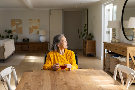 Senior Asian woman sitting in wheelchair at wooden table wearing mustard sweater holding teacup
