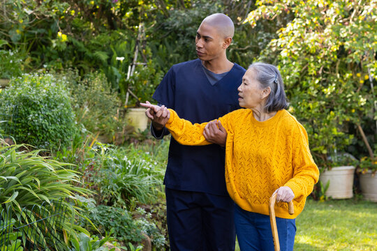 Senior Chinese woman in mustard sweater using cane, African navy-scrub caregiver guiding in garden
