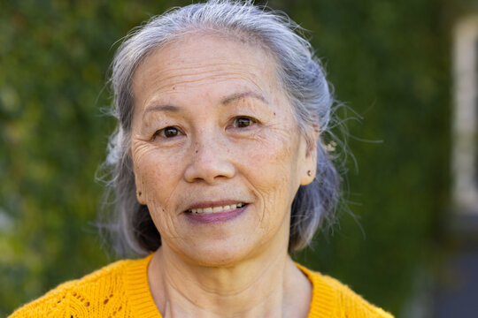 Senior Chinese woman smiling facing camera outdoors in garden wearing yellow knit sweater and studs