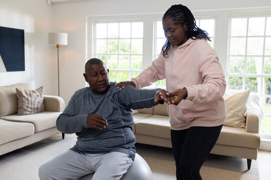 African American woman guiding older man sitting on silver ball, wearing pink hoodie in living room