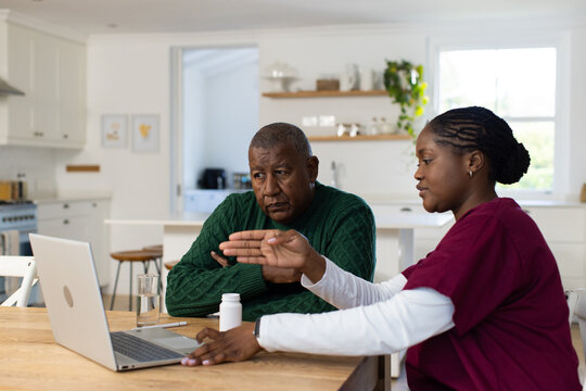 African American nurse in burgundy scrubs advising man at kitchen table with laptop and pill bottle