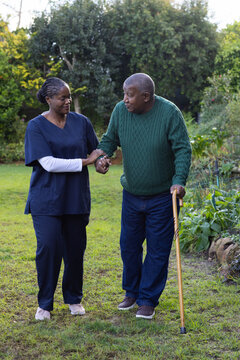 African American female caregiver in navy scrubs supporting senior man walking in yard with cane