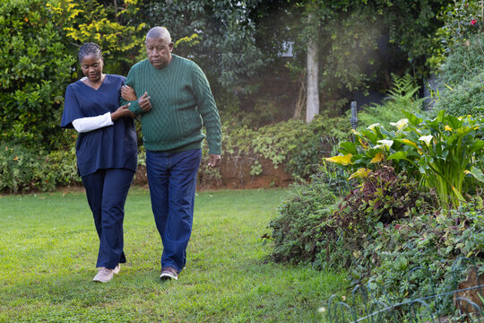 African American carer guiding mature adult man walking across garden with flowerbed in navy scrubs