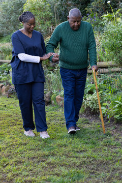 African American caregiver navy scrubs walking beside senior man on grassy path holding wooden cane