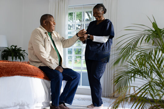 African American caregiver in scrubs smartwatch helping Senior man on bed in room with rust throw