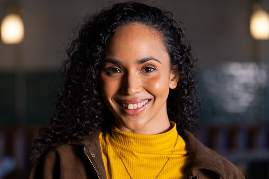 Woman with dark curly hair smiling, wearing yellow turtleneck and pendant necklace in cafe