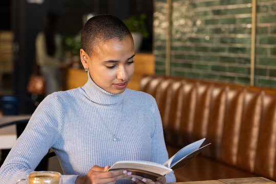 Woman with shaved head sitting in cafe booth reading paperback book and holding coffee cup