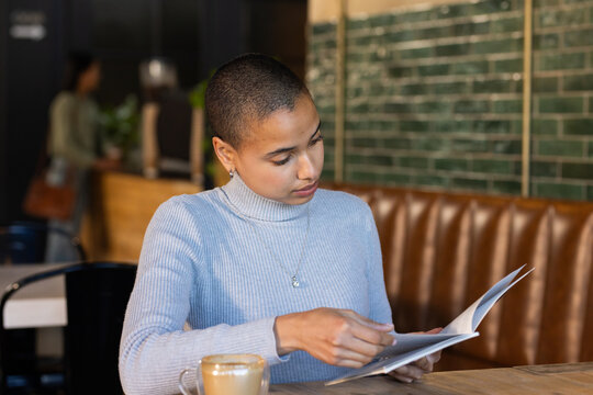 African American woman sitting at cafe table flipping booklet holding latte, wearing turtleneck