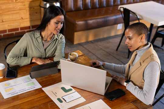 Diverse female coworkers meeting at cafe table reviewing laptop mockups, charts