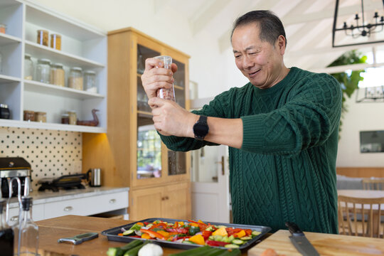 Senior Asian man grinding pepper over baking tray of chopped veggies on kitchen island, copy space
