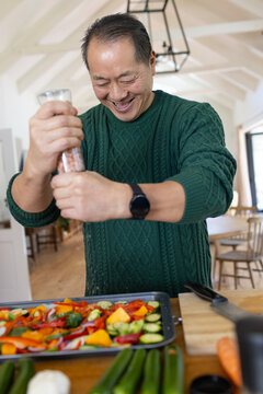 Mature Asian man seasoning veg on baking tray at counter using clear grinder in green sweater