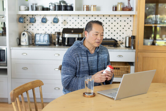 Senior Asian male reading pill bottle label at round wooden table in kitchen, viewing silver laptop