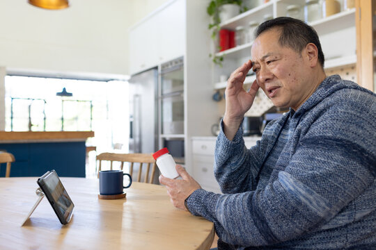 Senior man in blue hooded sweater sitting at kitchen table holding pill bottle and viewing tablet
