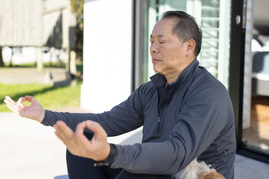 Senior Asian man meditating on patio near sliding glass door wearing dark zip jacket with dog
