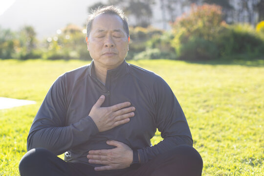 Asian man sitting cross-legged on lawn keeping hands on chest, abdomen wearing jacket, smartwatch