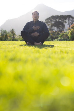 Mature Asian male meditating cross-legged on lawn, wearing dark jacket and pants
