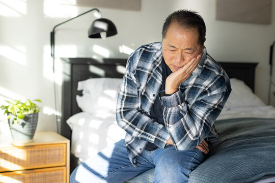 Senior Asian man sitting on bed, wearing plaid shirt, resting cheek on hand, sunlight streaming