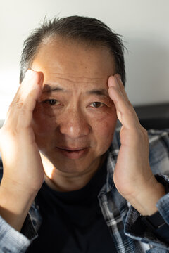 Mature Asian man pressing temples while sitting in room wearing checked flannel and wristwatch