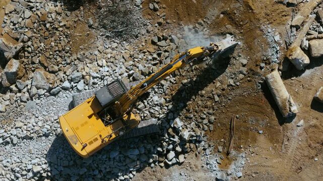 Aerial view Yellow excavator with a hydraulic breaker smashes boulders in a stone quarry, dust rising. Top down aerial of crawler tracks, crushed gray rock piles, brown dirt and long shadows