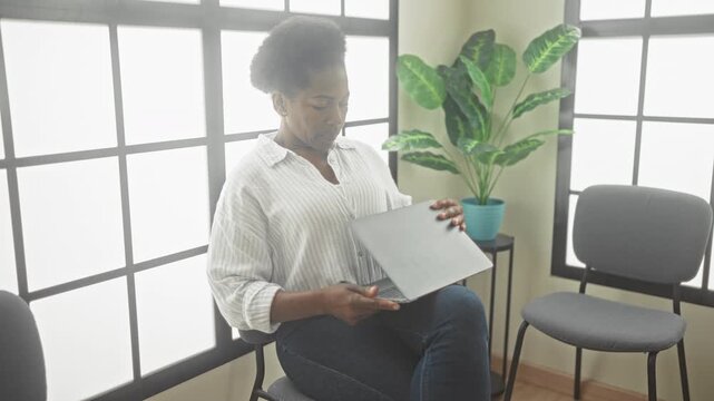Woman holding laptop while sitting on a gray chair beside a large grid window in a waiting room inside a building; anticipation.