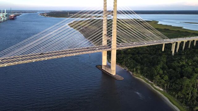 Drone moves laterally past bridge pylons beside a wooded shoreline in 4K video.

