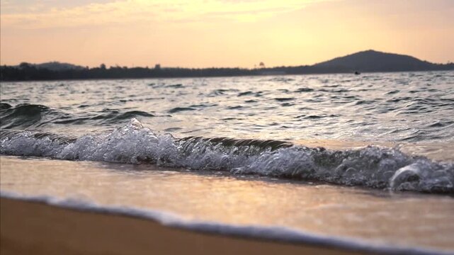 Sunset waves meet sandy shore in quiet coastal scene. Warm light shimmers across water as small breakers spread foam near beach, with dark hillside silhouetted against glowing evening sky.