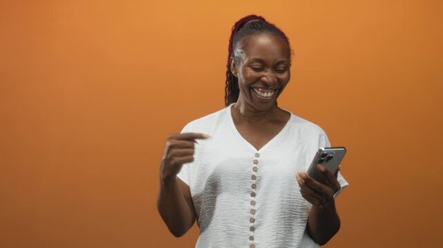 African american woman smiling while holding smartphone and gesturing with hand against orange studio backdrop; joyful connection.