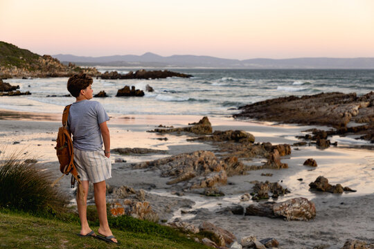 rear view of teen boy with backpack looking at rocks and ocean, Kammabaai Beach, Hermanus, South Africa