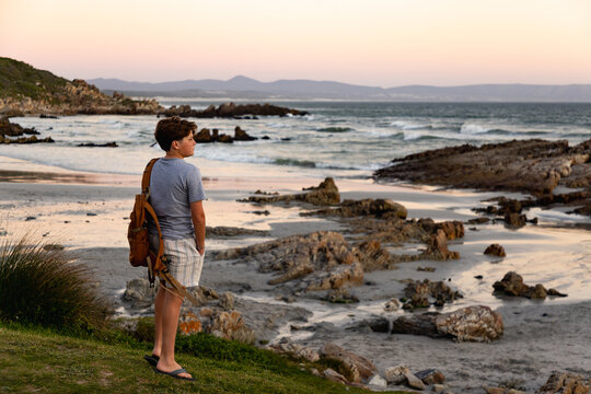 rear view of teen boy with backpack looking at rocks and ocean, Kammabaai Beach, Hermanus, South Africa