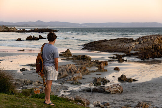 rear view of teen boy with backpack looking at rocks and ocean, Kammabaai Beach, Hermanus, South Africa