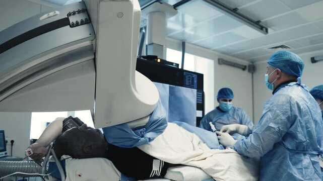 Patient lying under a C-arm system while doctors perform an angiography procedure under real-time X-ray guidance.
