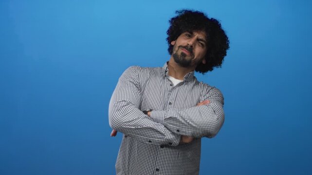 Man with curly hair giving double thumbs up and smiling in blue studio, wearing checked shirt and smartwatch; confidence friendly upbeat.