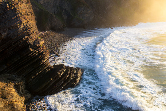 Geological flysch rock layers and ocean waves crashing on coastal cliff at sunset, Spain