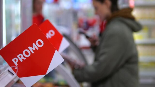 Shopper compares packaged goods beside prominent red promo tags in grocery aisle. Adult customer checks item near chilled shelves, considering discount during routine shopping and examining product