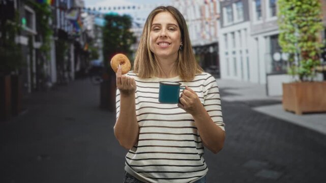 Young hispanic blonde woman holding a donut and a blue mug on a city street, lips puckered in a kiss gesture while smiling; simple joy.