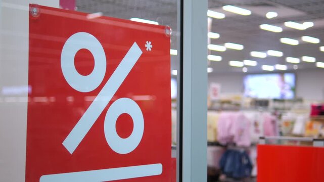 Red sale placard with oversized percent symbol stands before clothing displays in retail store. Discount sign remains fixed on glass while shoppers browse merchandise in bright shop interior.