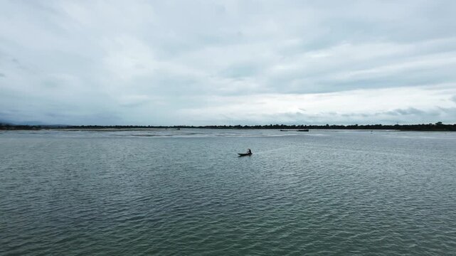 Aerial forward flight over lone boatman in small wooden boat on wide calm river under moody overcast sky