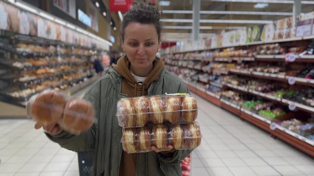 Woman selects packaged bread from supermarket shelves, carefully balancing several loaves in her arms. Focused shopper checks bakery goods and smiles as she finishes stacking them.