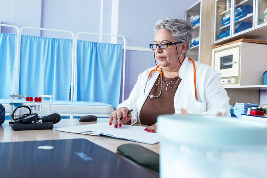 An older female doctor stands in her medical office, looking professional and confident. The portrait reflects her experience and dedication to caring for patients