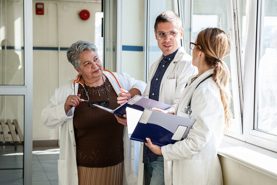 Three doctors are getting ready in the hospital hallway before heading to a meeting. They talk briefly and review their notes as they prepare to discuss their work together.