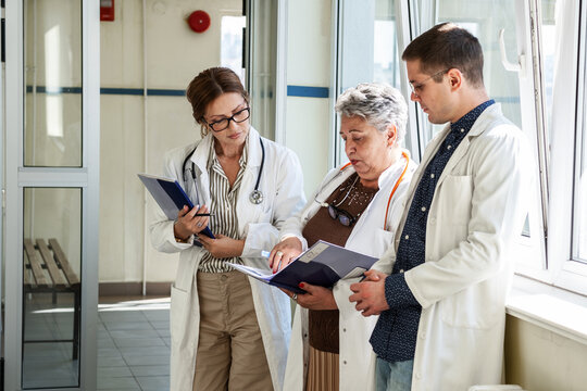 Three doctors are getting ready in the hospital hallway before heading to a meeting. They talk briefly and review their notes as they prepare to discuss their work together.