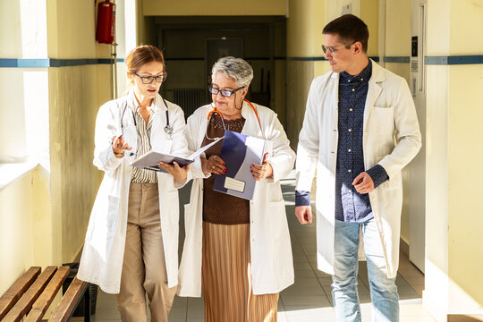 Three doctors are getting ready in the hospital hallway before heading to a meeting. They talk briefly and review their notes as they prepare to discuss their work together.